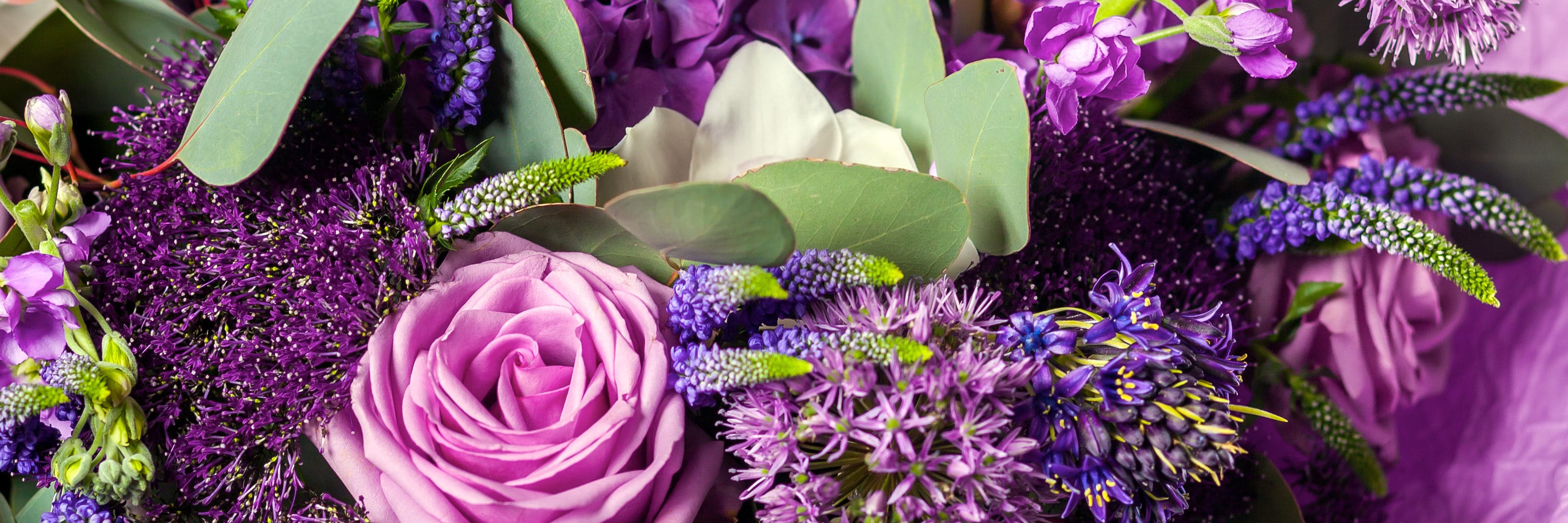 A bouquet of various purple, lavender, and white flowers, including roses and alliums, interspersed with green leaves; the flowers are densely packed, creating a vibrant, colorful arrangement.